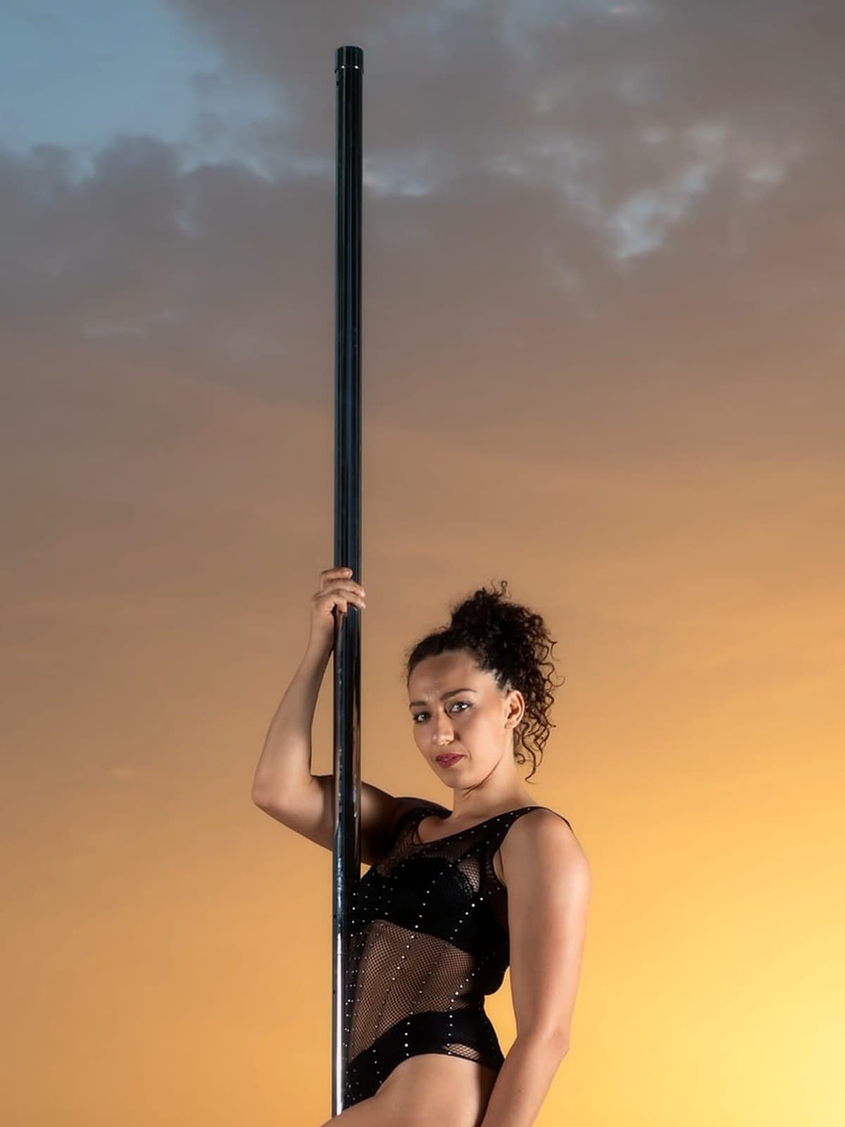 Sue en body noir et talons, à la pole au bord de la mer au coucher du soleil